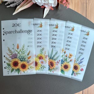 May include: A set of six transparent budget sheets with floral designs, including sunflowers and other flowers. Each sheet has the text "20€" and "Sparchallenge" printed on it. The sheets are hole-punched for binder use.
