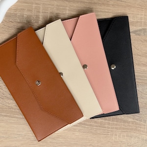Könnte beinhalten: Vier lederne Umschlag-Geldbörsen in Braun-, Creme-, Rosa- und Schwarztönen. Jede Geldbörse hat einen Klappenverschluss mit einem silbernen Druckknopf. Die Geldbörsen sind diagonal angeordnet.