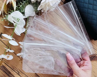 May include: Clear plastic binder pouches with zipper closures. The pouches are held in a hand, with a wooden surface in the background. Artificial flowers in shades of pink and white are also visible.