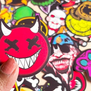 Può includere: Una collezione di adesivi colorati con vari disegni. Un adesivo presenta una faccia da diavolo rossa con corna bianche, occhi a croce neri e un ampio sorriso. Altri adesivi includono teschi, clown e personaggi dei cartoni animati.