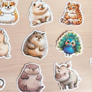 K&ouml;nnte beinhalten: Eine Sammlung farbenfroher Tieraufkleber, darunter ein Corgi, ein Leopard, ein Pinguin und ein Nashorn. Die Aufkleber sind im Aquarellstil mit wei&szlig;en R&auml;ndern gehalten und auf einer hellen Holzoberfl&auml;che angeordnet.