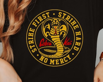 May include: Black t-shirt featuring a circular Cobra Kai design with a yellow cobra, red text, and the phrases "Strike First, Strike Hard, No Mercy." The design is centered on the front of the shirt.