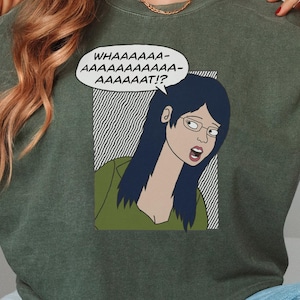 May include: An olive green t-shirt with a comic strip graphic. The graphic depicts a woman with blue hair and glasses, and a speech bubble that says "WHAAAAAAA- AAAAAAAAAA- AAAAAAAAT!?"