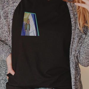 Puede incluir: Camiseta negra con un pequeño gráfico colorido de una persona en una puerta. La persona lleva una chaqueta verde y pantalones grises. La camiseta se combina con vaqueros azules y una rebeca gris.