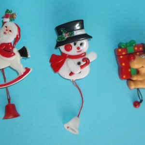 May include: Three Christmas-themed pins featuring a Santa Claus on a white rocking horse, a snowman wearing a black top hat and red scarf, and a brown reindeer holding a red gift box with a green ribbon, each with a small dangling bell attached.