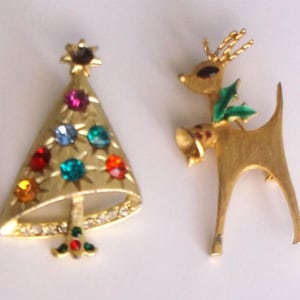 May include: Two vintage Christmas brooches. One is a gold-toned Christmas tree with colorful gemstone accents. The other is a gold-toned reindeer with green holly leaves and a red gemstone on its collar.