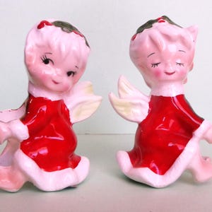 May include: Two vintage ceramic figurines of cherubic angels. Each angel is depicted in a seated position, wearing a red dress trimmed with white fur. The angels have pink skin, white wings, and dark green hair with red accents. The figurines are likely decorative items.