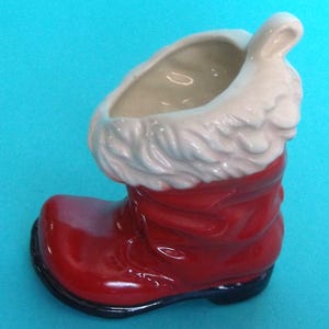 May include: A ceramic Santa boot planter, primarily red with a white fluffy trim and a black sole. The boot has a glossy finish and a hollow interior for planting small flowers or displaying holiday decorations. The background is a solid light blue.