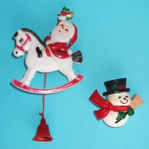 May include: Two holiday pins featuring a white rocking horse with a red-suited Santa Claus holding a gold bell, and a snowman wearing a black top hat with a red scarf and green mittens, holding a small wooden broom against a bright blue background.