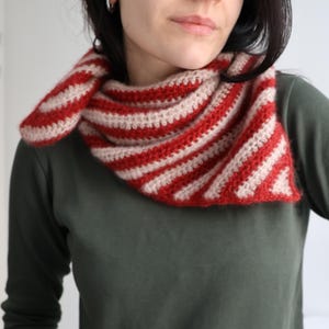May include: A red and cream-colored crocheted scarf. The scarf is striped with alternating bands of red and cream. The scarf is wrapped around the neck and shoulders. The material appears to be soft and warm.