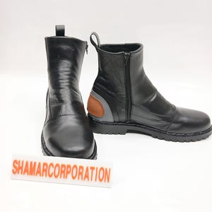 May include: Two black leather boots with zippers and pull tabs. The boots have a rounded toe and a small brown and gray accent on the side. The boots are on a white surface. The text "SHAMARCORPORATION" is visible.