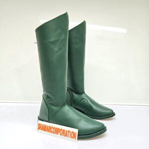 Puede incluir: Un par de botas altas de cuero verde. Las botas tienen una textura suave y una punta afilada. La caña de las botas es recta y llega hasta la mitad de la pantorrilla. Las botas se exhiben sobre una superficie blanca.