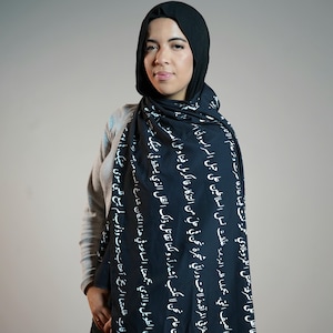 Puede incluir: Un hiyab y una bufanda negros con escritura árabe blanca. La bufanda está drapeada sobre los hombros. La mujer de la imagen lleva una camisa gris de manga larga.