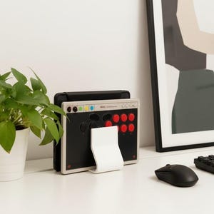 May include: A rectangular arcade controller featuring a black faceplate with red and black circular buttons, a light gray frame, and a white stand. The controller displays the text "Haute42" and "M16" on the top edge, with small colored circular indicators.