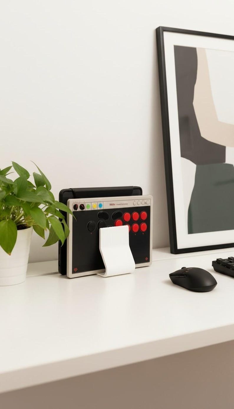 May include: A rectangular arcade controller featuring a black faceplate with red and black circular buttons, a light gray frame, and a white stand. The controller displays the text "Haute42" and "M16" on the top edge, with small colored circular indicators.