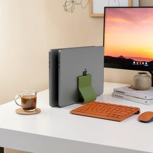 Könnte beinhalten: Ein dunkelgrauer Laptop, der von einem salbeigrünen, gerippten L-förmigen Ständer aufrecht gehalten wird. Das Set umfasst eine orangefarbene kabellose Tastatur, eine passende orangefarbene Computermaus, eine Kaffeetasse aus Glas und zwei Bücher.