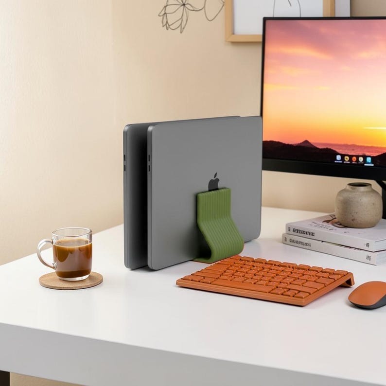 May include: A dark gray laptop held upright by a sage green, ribbed, L-shaped stand. The setup includes an orange wireless keyboard, a matching orange computer mouse, a glass mug filled with coffee on a coaster, and two stacked books.