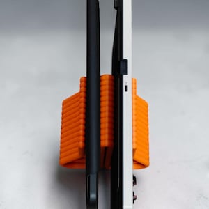 May include: An orange, ribbed, U-shaped stand holding two electronic devices upright. One device is encased in a slim black cover, while the other is a white tablet or handheld console featuring a visible USB-C port and small black buttons on the side.