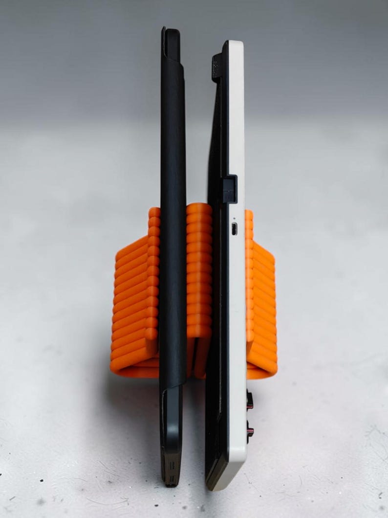 May include: An orange, ribbed, U-shaped stand holding two electronic devices upright. One device is encased in a slim black cover, while the other is a white tablet or handheld console featuring a visible USB-C port and small black buttons on the side.