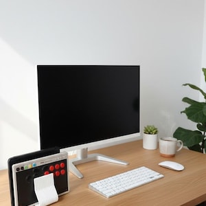 May include: A computer monitor with a black screen, a white wireless keyboard, a white wireless mouse, and a black game controller with red and yellow buttons resting in a white ridged stand on a light wood desk surface.
