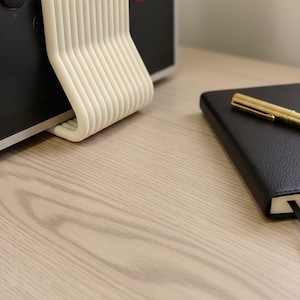 May include: A cream-colored, U-shaped plastic stand with multiple vertical ribs, positioned against a black arcade controller featuring red and black circular buttons labeled P1 and P3. A black notebook with a gold pen rests nearby.