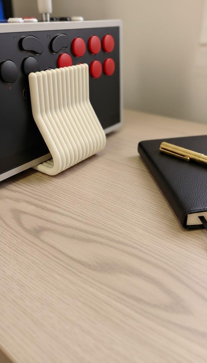 May include: A cream-colored, U-shaped plastic stand with multiple vertical ribs, positioned against a black arcade controller featuring red and black circular buttons labeled P1 and P3. A black notebook with a gold pen rests nearby.