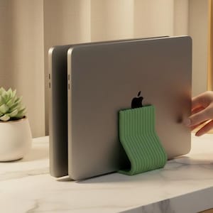 May include: A sage green, ribbed, U-shaped vertical laptop stand holding two silver laptops in an upright position. The stand features a textured, parallel-line design and provides a stable base for storing electronic devices vertically on a desk.