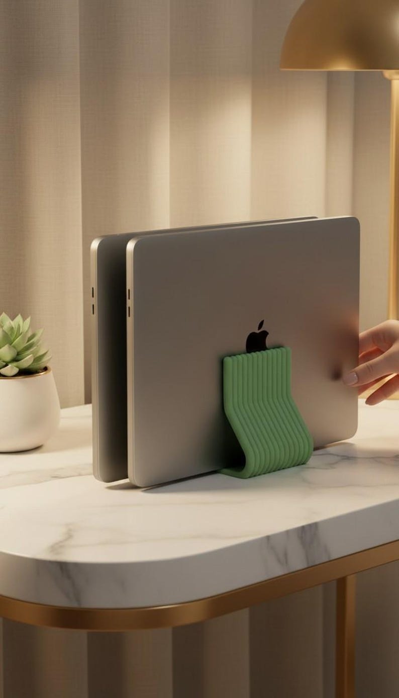 May include: A sage green, ribbed, U-shaped vertical laptop stand holding two silver laptops in an upright position. The stand features a textured, parallel-line design and provides a stable base for storing electronic devices vertically on a desk.