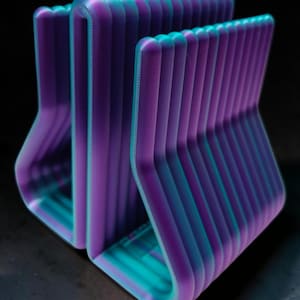 May include: A 3D printed desk organizer featuring a series of parallel, curved vertical dividers. The object displays a vibrant gradient color transition, shifting from deep purple on the outer edges to a bright teal blue in the center.