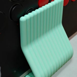 May include: A mint green, ribbed, L-shaped support stand positioned against a black arcade controller panel. The stand features a series of vertical, rounded ridges and a smooth, curved base that provides stability against the flat surface.