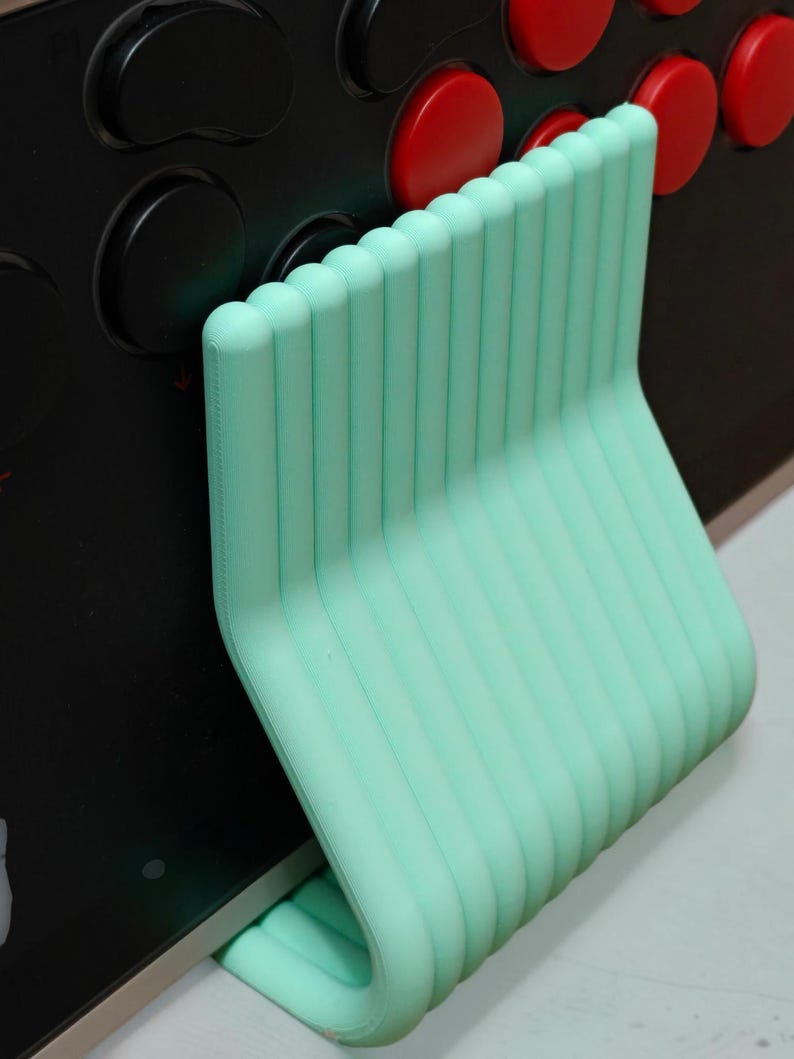 May include: A mint green, ribbed, L-shaped support stand positioned against a black arcade controller panel. The stand features a series of vertical, rounded ridges and a smooth, curved base that provides stability against the flat surface.