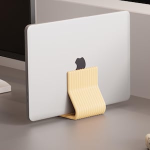 Op de afbeelding: Een lichtgele, geribbelde, U-vormige laptopstandaard die een zilveren laptop in een verticale positie houdt. De standaard is voorzien van een reeks parallelle ribbels langs het buitenoppervlak voor een stabiele basis op een bureau.
