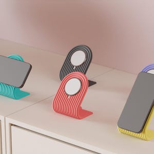 May include: A collection of colorful phone stands in teal, coral, black, and yellow with a purple accent. Each stand has a ribbed design and holds a gray smartphone. The stands are displayed on a white surface.