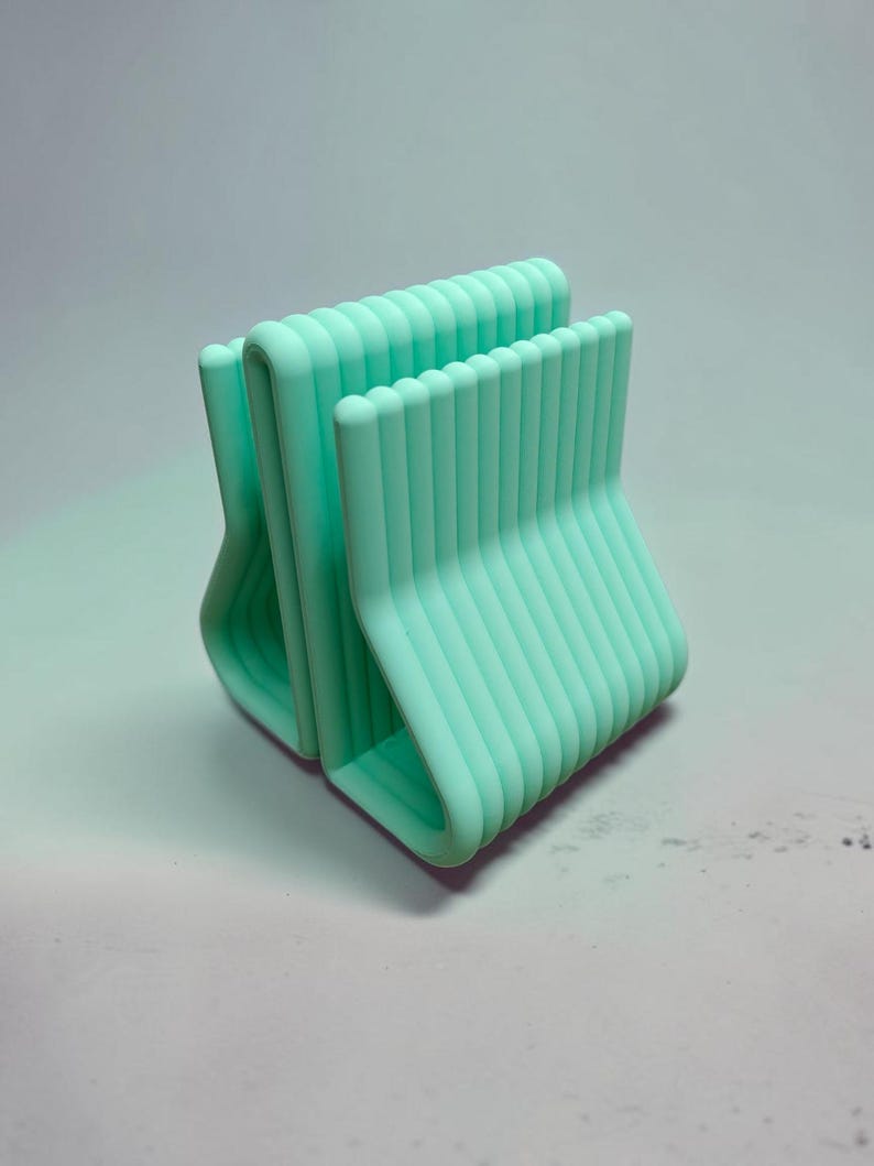 May include: A mint green desk organizer featuring a series of parallel, vertical, rounded bars arranged in a repeating, stepped pattern. The structure is composed of multiple connected U-shaped segments that create a ribbed, textured appearance.