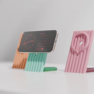 May include: Three colorful phone stands in orange, mint green, and pink. The mint green stand holds a phone displaying a clock and calendar. The pink stand has a circular cutout. All stands have a ribbed design.
