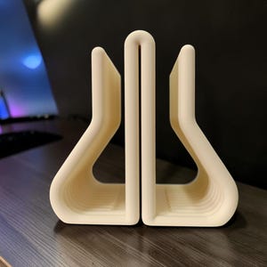 May include: A cream-colored, U-shaped desk organizer featuring a central vertical divider and two symmetrical side compartments with curved, rounded edges. The object has a smooth, matte finish and a structured, open-frame design.