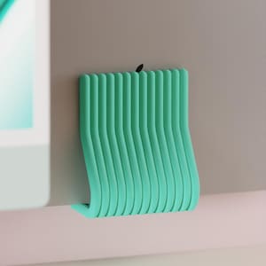 Könnte beinhalten: Ein türkisfarbener, wellenförmiger Kabelorganizer aus flexiblem Material. Der Organizer ist an einer grauen Oberfläche befestigt und dient zum Halten und Verwalten von Kabeln. Das Design ist minimalistisch und modern, geeignet für den Heim- oder Bürogebrauch.