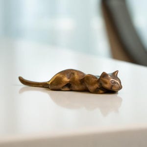 Puede incluir: Una pequeña figurita de metal dorada de un gato acostado sobre una superficie blanca.