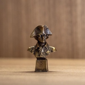 Pode incluir: Um busto de cor bronze de uma figura histórica, possivelmente um líder militar, usando um chapéu bicorne e um casaco com gola. A escultura é montada em uma pequena base retangular. O fundo é uma superfície de madeira.