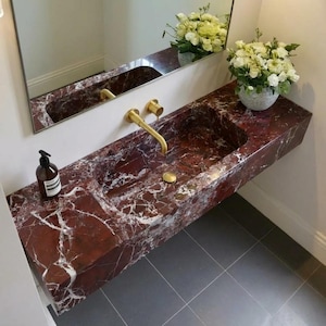 Può includere: Un moderno lavabo da bagno in marmo rosso con venature bianche. Il lavabo ha una forma rettangolare con due vasche e un rubinetto dorato. Uno specchio è appeso sopra il lavabo. Una bottiglia di sapone e un vaso di fiori sono sul piano.