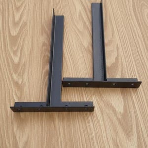May include: Two black metal shelf brackets, each with a vertical support and a horizontal base with pre-drilled holes for mounting. The brackets are designed for supporting shelves and are shown against a light wood grain background.