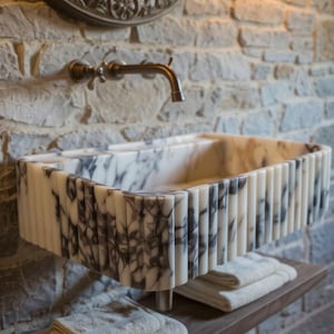 May include: A rectangular marble sink with a fluted design, featuring a mix of white and grey veining. The sink is mounted on a wooden shelf with folded towels, set against a stone wall with a bronze tap.
