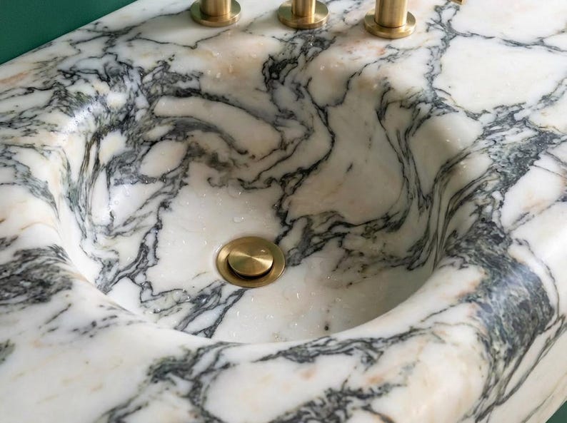 May include: A close-up of a marble sink with a round basin and a gold-colored drain. The sink has a white background with gray and black veining. Three gold-colored faucets are visible in the background.