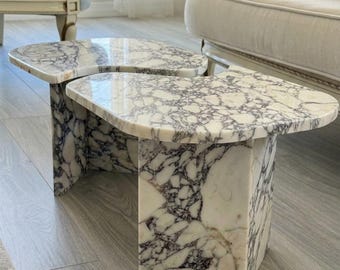 May include: Two marble side tables with a white and purple veined pattern. The tables have a rectangular base and a rounded top. The tables are made of marble and are placed on a light wood floor.