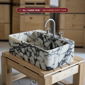 May include: A rectangular white marble sink with prominent black veining, featuring a matte silver faucet and two matching cross-handle knobs. The sink is positioned on a wooden pallet. Text above reads: ALL TAXES PAID • DELIVERED DUTY PAID.