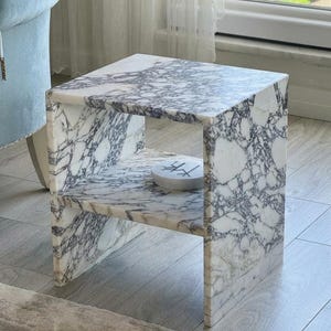 Puede incluir: Una mesa auxiliar cuadrada de mármol con un estante. La mesa tiene un patrón veteado blanco y gris. Un objeto pequeño, redondo y blanco con marcas negras se encuentra en el estante. La mesa es de piedra y tiene un diseño moderno.