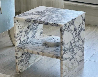 May include: A square marble side table with a shelf. The table has a white and gray veined pattern. A small, round, white object with black markings sits on the shelf. The table is made of stone and has a modern design.