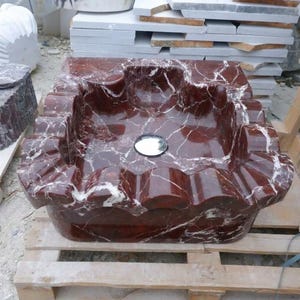 May include: A red marble sink with white veining. The sink has a unique, scalloped edge design and a circular drain. The sink is made of polished stone and sits on a wooden pallet.
