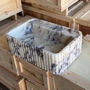 Könnte beinhalten: Ein weißes Marmorspülbecken mit einem gerippten Design und schwarzen Adern. Das Spülbecken steht auf einer Holzkiste.