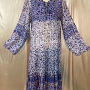 Puede incluir: Un vestido largo y fluido con un estampado de cachemira en tonos azules, morados y blancos. El vestido presenta mangas largas, un escote en V con cordón y una falda escalonada. La tela parece ligera y transparente.
